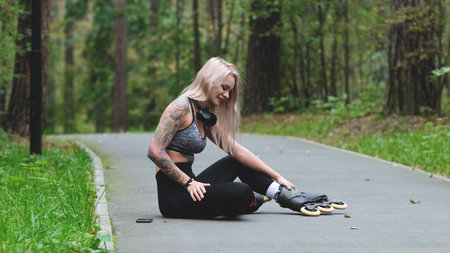 Attractive blonde relaxes while roller skating. She sits on the bike path and poses for the camera. Stunning Caucasian woman with a beautiful figure uses roller skates for outdoor sports. The girl feels free and easy while roller skating alone in the city park.の写真素材