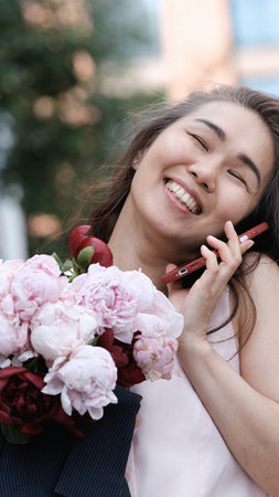Beautiful woman with bouquet of peonies talking on phone and looking happy outdoors. Urban lifestyle concept.の写真素材