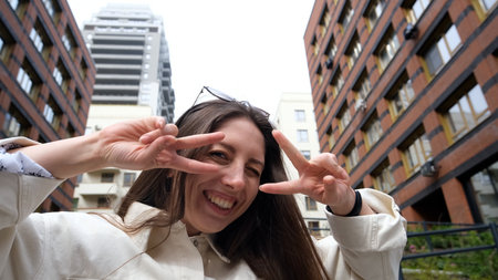 Cute cheerful charming girl poses for camera in summer on city street, hands and fingers showing V...