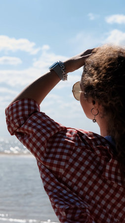 Young woman with curly hair in red checkered shirt walking along seashore enjoying summer holidays. Vacation, summer, weekendの写真素材
