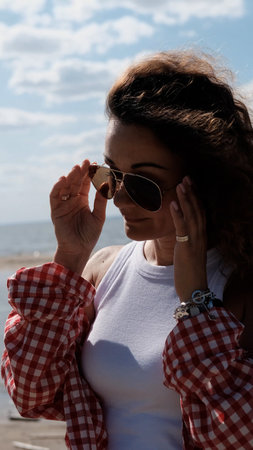 Young woman with curly hair in red checkered shirt walking along seashore enjoying summer holidays....