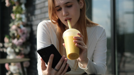A young girl uses a smart phone and drinks a mango or orange juice cocktail while sitting at a table...
