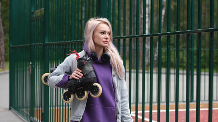 Blonde woman walking through city park holding roller skates enjoying beautiful dayの写真素材