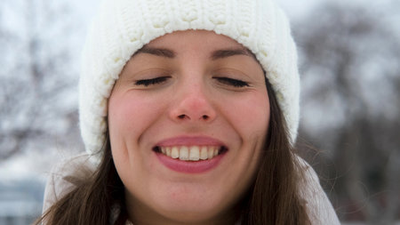 Portrait of a woman in a warm white knitted hat outdoors in winter. Young woman enjoying the beautiful weather and first snow. Active winter recreation. Wide, beautiful smile. Close-up.の写真素材