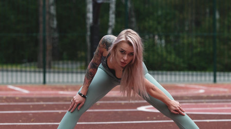 Young blonde woman in sportswear doing stretching exercises standing on basketball court in the morning. Woman feels pleasantly tired after intense daily workout program. Genuine candid photoの写真素材