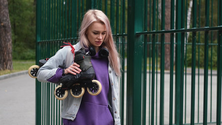 Blonde woman walking through city park holding roller skates enjoying beautiful day