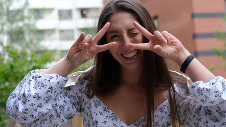 Cute cheerful charming girl poses for camera in summer on city street, hands and fingers showing V signの写真素材