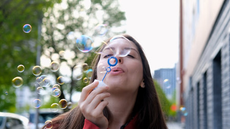 Young beautiful woman in burgundy coat blowing soap bubbles on city street. Freedom concept