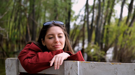 Portrait photo of beautiful young smiling woman in burgundy coat sitting on bench outdoors in park.の写真素材