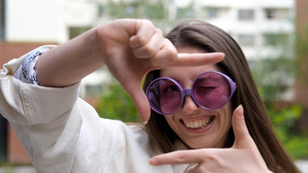 A sweet, cheerful, and charming girl poses for the camera on a city street in the summer. The woman holds up a hand-made frame, like a photograph. A photo frame made by a young hipster girl.の写真素材