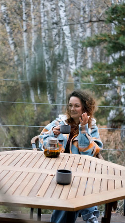 A woman with curly hair drinks tea and relaxes on the terrace of her house on an autumn day.の写真素材