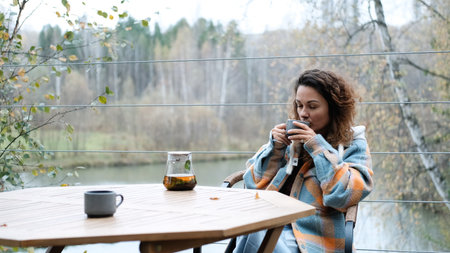 A sweet woman with curly hair drinks tea and relaxes on the terrace of her house on an autumn day. Health care, sincerity, a sense of balance and calm.の写真素材