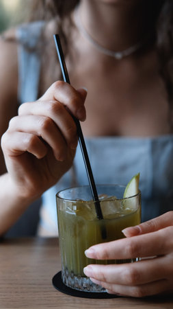 A close-up of a sweet young woman with long curly hair drinking a cocktail through a straw at a city bar. Beautiful female lips take a sip from a glass using a straw at a cafe. A brunette samples a delicious drinkの写真素材