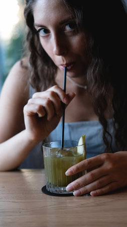 A close-up of a sweet young woman with long curly hair drinking a cocktail through a straw at a city bar. Beautiful female lips take a sip from a glass using a straw at a cafe. A brunette samples a delicious drinkの写真素材