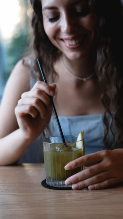 A close-up of a sweet young woman with long curly hair drinking a cocktail through a straw at a city bar. Beautiful female lips take a sip from a glass using a straw at a cafe. A brunette samples a delicious drinkの写真素材
