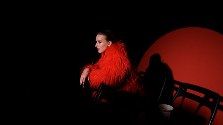 Portrait of a beautiful young woman in a red fur coat against a red background, posing for a cinema-style photo. The woman smiles beautifully, sitting on chairs in the light of a projector during a movie screeningの写真素材