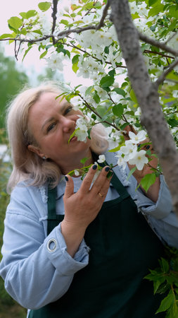 A portrait of a happy woman gardener, standing next to a blossoming apple tree, smiling, enjoying...