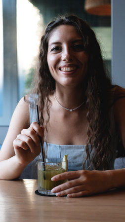 A close-up of a young woman with long curly hair drinking a cocktail through a straw at a city bar....