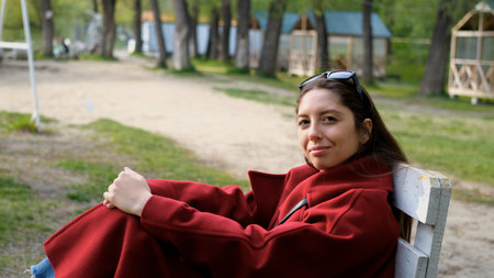 Portrait photo of beautiful young smiling woman in burgundy coat sitting on bench outdoors in park.の写真素材