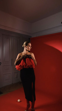 Portrait of a beautiful young woman in a red fur coat against a red background, posing for a photo. The woman smiles beautifully, sitting on chairs in the light of a projector during a screeningの写真素材