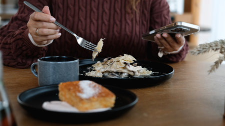 A woman in a brown sweater eats pasta in a restaurant and texts on her smartphone. Close-up of the...