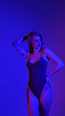 A blonde with a beautiful figure poses in a black bodysuit in a studio. Studio lighting, low exposure, monochrome blue light.の写真素材