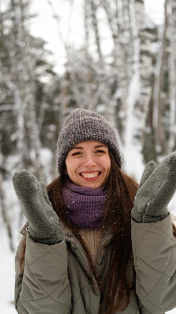 A beautiful young woman in winter clothes is happily spending her free time in a snowy forest. She...