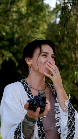 A beautiful brunette in a bohemian style relaxes outdoors in summer against a backdrop of a green forest, holding a large bunch of juicy black grapesの写真素材