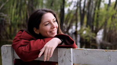 Portrait photo of beautiful young smiling woman in burgundy coat sitting on bench outdoors in park.の写真素材