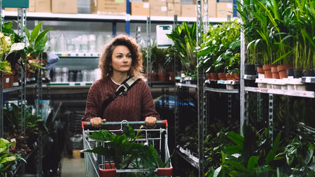 A happy woman with curly hair buys potted flowers at a garden center. The woman carries the flowers in a shopping cart.