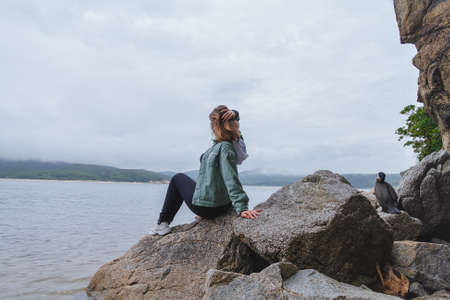 Young beautiful woman sits on the rocks on the seashore and looks into the distance.