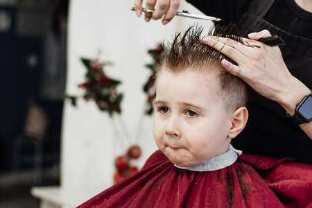 Little boy gets his hair cut with a pair of barber scissors. Close-up of woman hands grooming kid boy hair in barber shop. Portrait of male child at the barber shop to cut his hair.の写真素材
