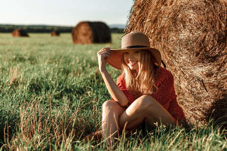 Happy young woman resting near a bale of hay in a summer field at sunset. Young blonde woman in a straw hat sits by a haystack and smile. An atmospheric calm moment. Rural slow lifeの写真素材
