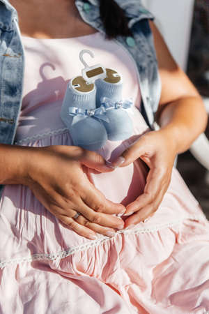 A pregnant woman in a pink dress holds her heart-shaped hands on her stomach. Blue booties on a...