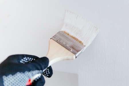 Close-up of a workers hand with a brush painting a wall with white paint.