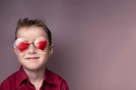 Smiling little boy in glasses with red hearts and a burgundy shirt, pink background. Valentine's...