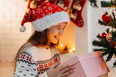 Girl in a red Santa hat opens a gift at home in the living roomの写真素材