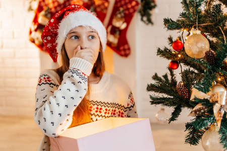 Girl in a red Santa hat opens a gift at home in the living roomの写真素材