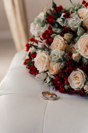 Gold wedding rings next to the bride's bouquet of beige roses with red berries lying on a white leather chair, close-upの写真素材