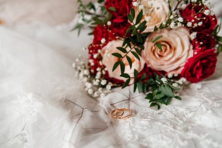 Gold wedding rings near the bride's bouquet of beige-red roses, lying on the bride's white veil, close-upの写真素材