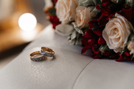 Gold wedding rings next to the bride's bouquet of beige roses with red berries lying on a white leather chair, close-upの写真素材