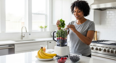 A young African woman with curly hair prepares a smoothie in a modern kitchen. She adds spinach to a blender filled with fruits. Bananas and berries are on the counter.の素材