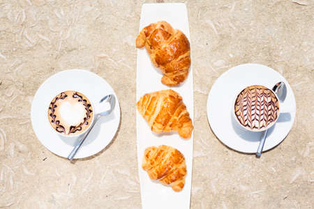Overhead view of cafe latte with topping on it. Standing on sand with croissants on white plate between cups. Horizontalの写真素材