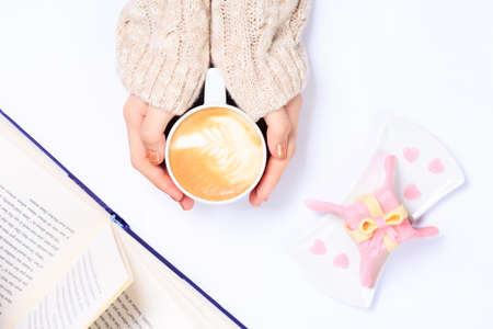 Female hands in beige knit holding cup of cappuccino. Gift alike dessert with heart shape sprinkles. Opened book. Flat lay. Valentines concept. Horizontalの写真素材
