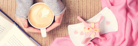 Girl holding cup of coffee with latte art. Pink textile, gift style cake, open book. Valentines day and leasure time concept. Pastel colors, warm toning. Horizontal, banner formatの写真素材