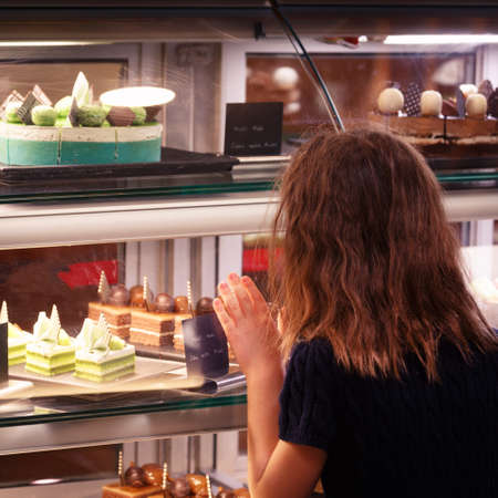 Little girl in confectionary shop touching the display. Sweet treats variety. Small business and child concept. No face visible. Square Warm white balanceの写真素材