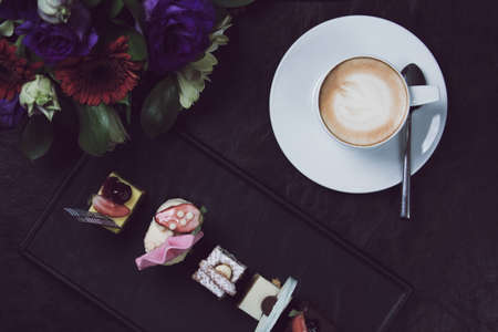 Coffee cup with latte art and flower bouquet. Concept of business event refreshment break. Top view. Mini desserts collection. Black background. Horizontalの写真素材