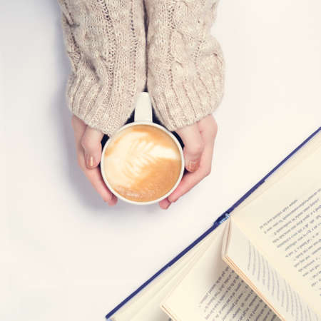 Girl holding cup of coffee with latte art. Open book. Leasure time concept. Pastel colors, warm...