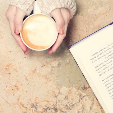 Girl holding cup of coffee with latte art. Open book, marble background. Leasure time concept. Pastel colors, warm toning. Squareの写真素材