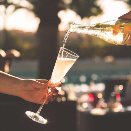 Waiter is pourring sparkling wine into a woman glass at the outdoor party.  Celebration concept. Event, party, wedding background. Toned image. Squareの写真素材